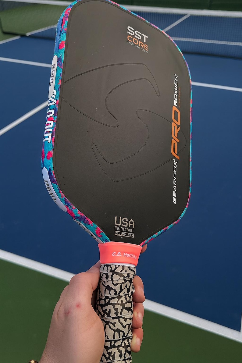 May include: A black pickleball paddle with a matte surface, featuring the text SST CORE, GEARBOX PRO POWER, and USA PICKLEBALL APPROVED. The edge guard has a vibrant blue and pink splatter pattern, and the handle has a grey and black elephant print.