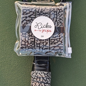 May include: A set of three grey and black elephant print overgrips packaged in a clear plastic pouch. A circular white label on the front features the text Kicks grips and the word DRY. One overgrip is wrapped around a black tennis racket handle.