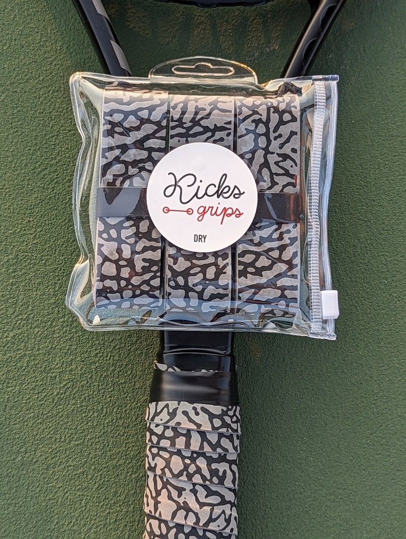 May include: A set of three grey and black elephant print overgrips packaged in a clear plastic pouch. A circular white label on the front features the text Kicks grips and the word DRY. One overgrip is wrapped around a black tennis racket handle.