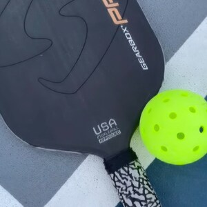 May include: A black Gearbox Pro Power elongated pickleball paddle with SST Core technology and a USA Pickleball Approved logo, paired with a bright neon yellow perforated pickleball. The paddle handle features a black and white textured pattern.