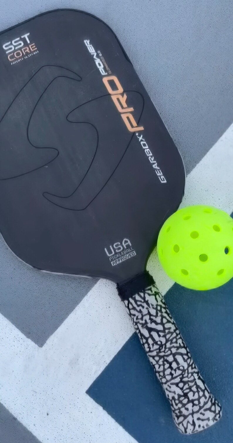 May include: A black Gearbox Pro Power elongated pickleball paddle with SST Core technology and a USA Pickleball Approved logo, paired with a bright neon yellow perforated pickleball. The paddle handle features a black and white textured pattern.