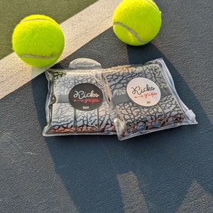 May include: Two clear plastic packages containing grey and black elephant print overgrips, labeled Kicks Grips Tacky and Kicks Grips Dry, positioned between two bright neon yellow tennis balls on a dark grey court surface.