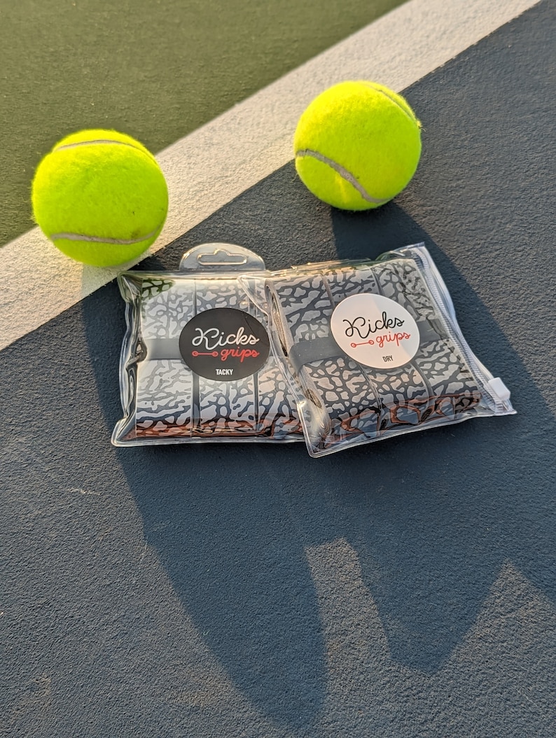 May include: Two clear plastic packages containing grey and black elephant print overgrips, labeled Kicks Grips Tacky and Kicks Grips Dry, positioned between two bright neon yellow tennis balls on a dark grey court surface.