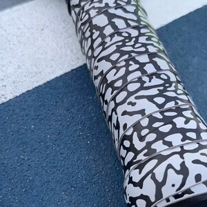 May include: A black pickleball paddle with a white and black elephant print grip. The paddle head features the text USA PICKLEBALL APPROVED. A bright yellow pickleball is partially visible at the top edge of the frame, resting near the paddle.