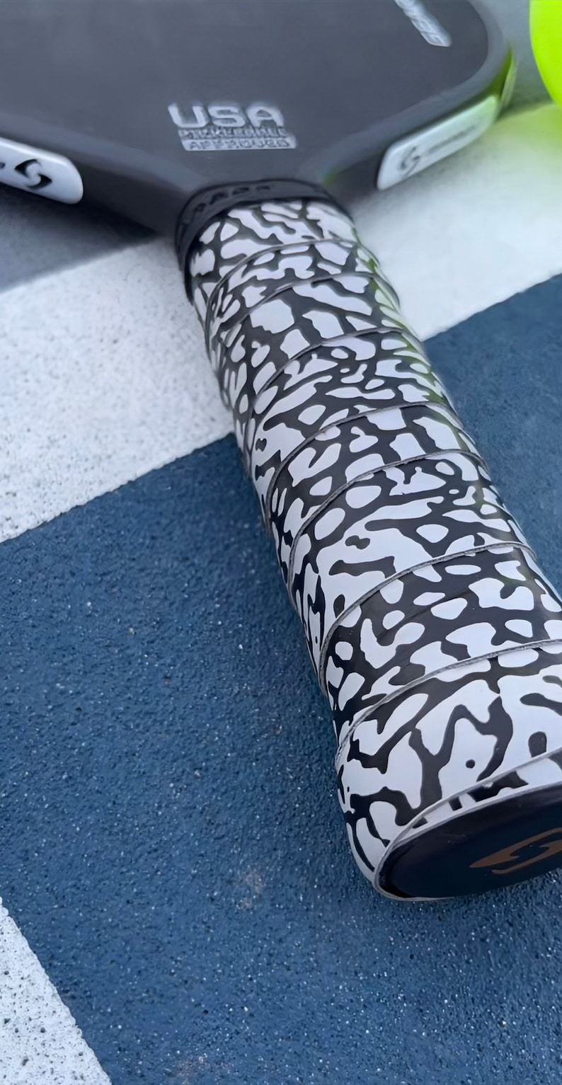 May include: A black pickleball paddle with a white and black elephant print grip. The paddle head features the text USA PICKLEBALL APPROVED. A bright yellow pickleball is partially visible at the top edge of the frame, resting near the paddle.