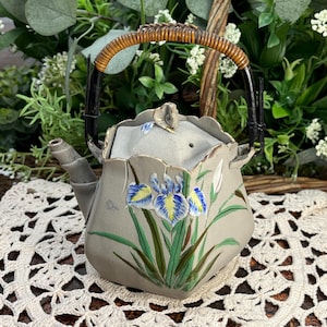 May include: A gray ceramic teapot with a woven handle and a floral design featuring blue, yellow, and green irises. The teapot has a lid with a small knob on top.