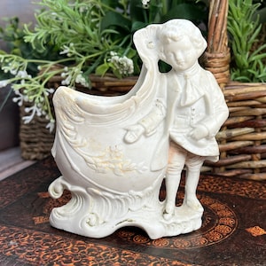 May include: White porcelain figurine of a man holding a decorative vase. The vase has a floral design and the man is wearing a jacket and pants. The figurine is standing on a brown surface.