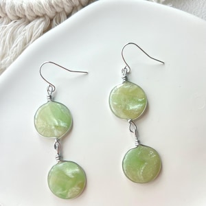 May include: Dangle earrings with two light green, circular pendants linked by silver wire. The earrings have silver-coloured ear hooks. The pendants have a marbled effect with slight colour variations. Each pendant is approximately 1.5 cm in diameter.