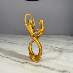 May include: A gold-colored abstract sculpture of two figures intertwined in a loving embrace. The sculpture is made of a smooth, shiny material and has a modern, minimalist design.