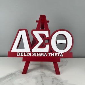 May include: A red and white Delta Sigma Theta sign displayed on a small easel. The sign features the Greek letters Delta, Sigma, and Theta in white, with the words "DELTA SIGMA THETA" printed below in white.