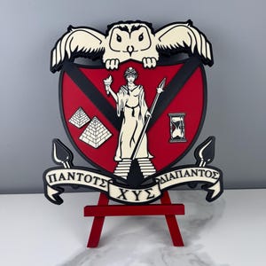 May include: A wooden crest featuring a red shield with a black border, an owl, and Greek text. The crest includes a white figure holding a torch and spear, two pyramids, and an hourglass. The crest is displayed on a red easel.