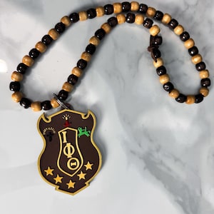 May include: A wooden bead necklace with a brown shield pendant. The pendant has gold trim, stars, and symbols. The beads alternate between light and dark brown.