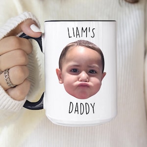 以下が含まれることがあります： 黒いハンドルと縁が付いた白いセラミックマグ。マグには、写真の上に「LIAM'S」、下に「DADDY」という文字が入った赤ちゃんの顔写真が写っています。白いセーターを着た人がマグを持っています。