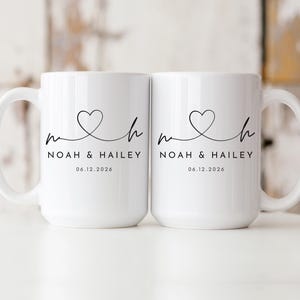 May include: Two white ceramic mugs with a heart design and the names "Noah & Hailey" printed in black. The date "06.12.2026" is printed below the names. Each mug has a large handle.