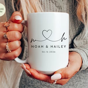 May include: A white 15oz ceramic mug with the text "NOAH & HAILEY" and a heart design. The date "06.12.2026" is printed below the names. The mug is held by a person.