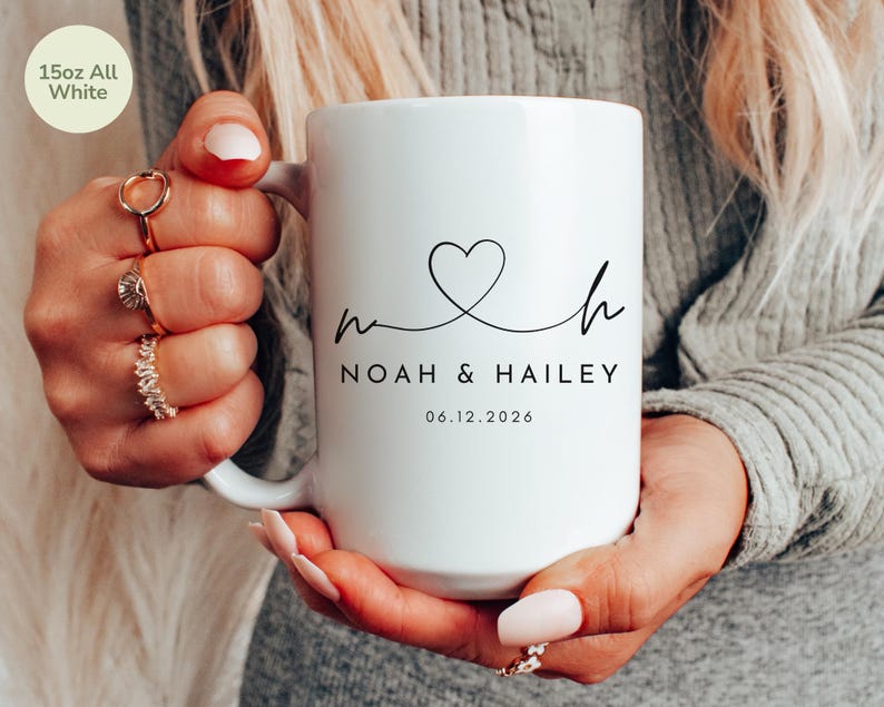 May include: A white 15oz ceramic mug with the text "NOAH & HAILEY" and a heart design. The date "06.12.2026" is printed below the names. The mug is held by a person.