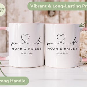 May include: Two white ceramic mugs with pink handles and interior rims. Each mug features a black heart design with the names "Noah & Hailey" and the date "06.12.2026". The mugs have a strong handle and a vibrant, long-lasting print.