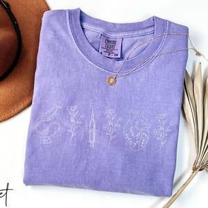 May include: A purple t-shirt with a white outline of a stethoscope, syringe, heart, and flowers. The shirt has the text "Comfort Colors" on the tag.