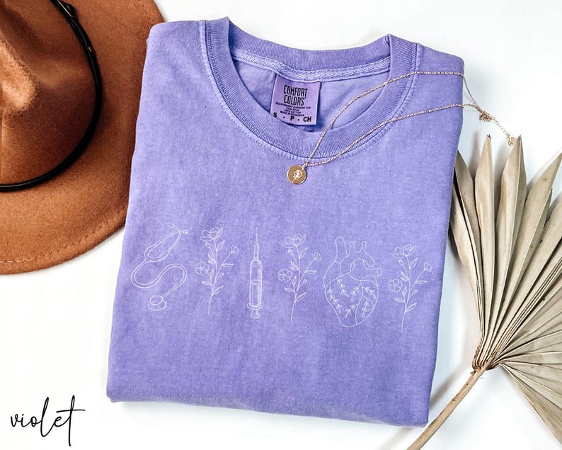 May include: A purple t-shirt with a white outline of a stethoscope, syringe, heart, and flowers. The shirt has the text "Comfort Colors" on the tag.