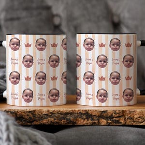 Może przedstawiać: Dwa ceramiczne kubki z czarnymi uchwytami i obw&oacute;dkami. Kubki mają powtarzający się wz&oacute;r twarzy dziecka, koron i pionowych pask&oacute;w w kolorze brzoskwiniowym i białym. Na kubkach wydrukowano r&oacute;wnież tekst "Liam's Papa".