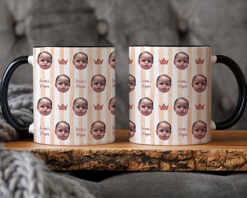 Może przedstawiać: Dwa ceramiczne kubki z czarnymi uchwytami i obw&oacute;dkami. Kubki mają powtarzający się wz&oacute;r twarzy dziecka, koron i pionowych pask&oacute;w w kolorze brzoskwiniowym i białym. Na kubkach wydrukowano r&oacute;wnież tekst "Liam's Papa".