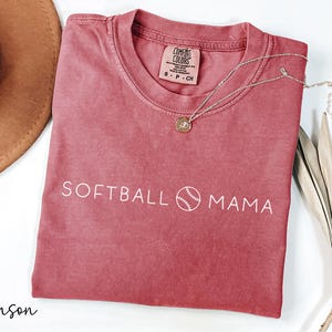May include: Crimson red t-shirt with the text "SOFTBALL MAMA" and a white baseball graphic. The shirt is made of a soft, comfortable fabric.