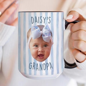 K&ouml;nnte beinhalten: Wei&szlig;er Keramikbecher mit schwarzem Henkel und Rand, mit dem Gesicht eines Babys mit blauer Schleife und dem Text "DAISY'S GRANDPA" auf einem blau-wei&szlig; gestreiften Hintergrund. Der Becher wird von einer Person gehalten.