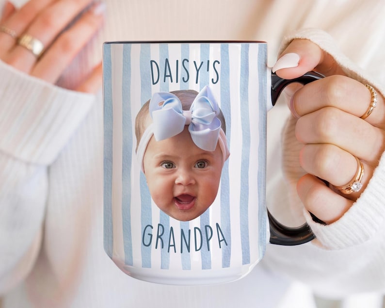 K&ouml;nnte beinhalten: Wei&szlig;er Keramikbecher mit schwarzem Henkel und Rand, mit dem Gesicht eines Babys mit blauer Schleife und dem Text "DAISY'S GRANDPA" auf einem blau-wei&szlig; gestreiften Hintergrund. Der Becher wird von einer Person gehalten.