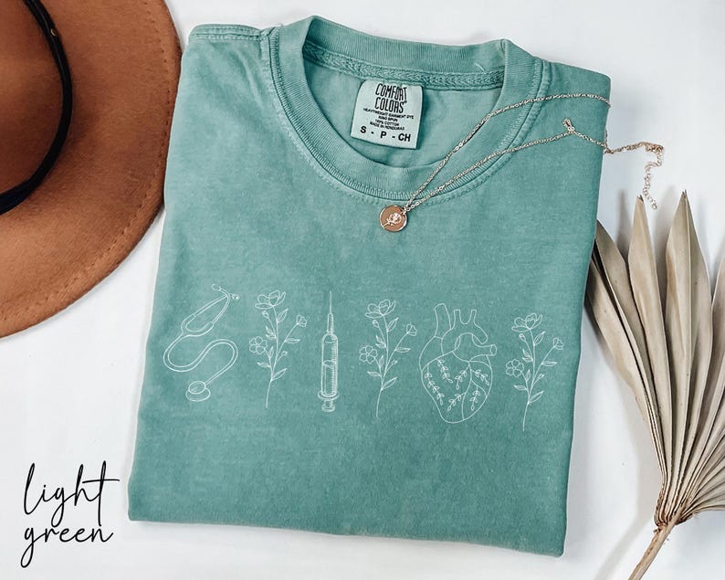May include: Light green t-shirt with a white outline of a stethoscope, syringe, heart, and flowers. The shirt has the text "Comfort Colors" and "S - P - CM" printed on the tag.