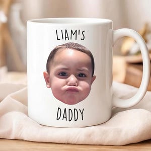 以下が含まれることがあります： 白いセラミックマグカップ、持ち手付き。マグカップには、子供の顔写真と、画像の上に「LIAM'S」、下に「DADDY」と黒いフォントで書かれた文字がプリントされています。マグカップはベージュ色の布の上に置かれています。