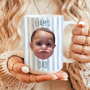 K&ouml;nnte beinhalten: Wei&szlig;er Keramikbecher mit blau-wei&szlig;en vertikalen Streifen. Der Becher zeigt ein Foto des Gesichts eines Kindes und die Worte "LIAM'S DAD" in blauer Schrift. Der Becher wird von einer Person gehalten, die goldene Ringe tr&auml;gt.