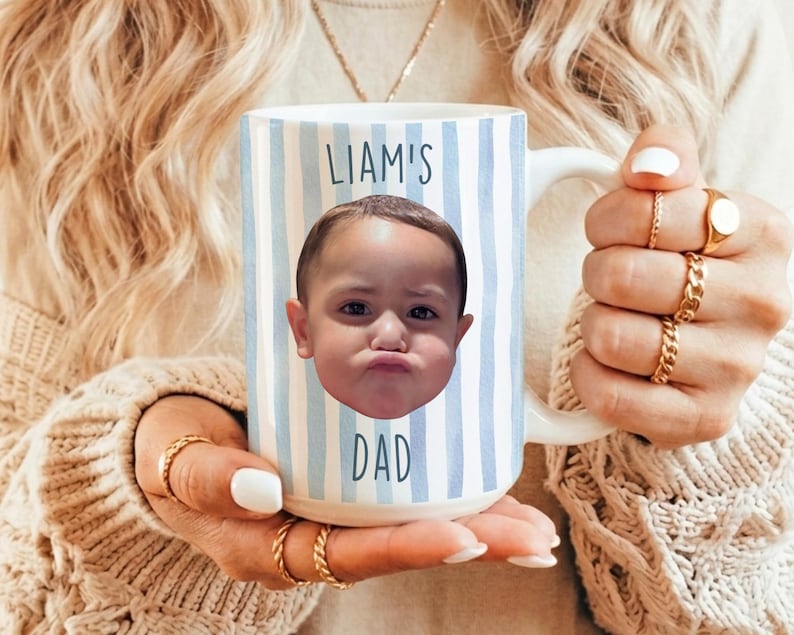 K&ouml;nnte beinhalten: Wei&szlig;er Keramikbecher mit blau-wei&szlig;en vertikalen Streifen. Der Becher zeigt ein Foto des Gesichts eines Kindes und die Worte "LIAM'S DAD" in blauer Schrift. Der Becher wird von einer Person gehalten, die goldene Ringe tr&auml;gt.