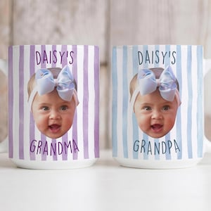 K&ouml;nnte beinhalten: Zwei wei&szlig;e Keramikbecher mit einem Babygesicht und den Texten "DAISY'S GRANDMA" und "DAISY'S GRANDPA". Die Becher haben vertikale Streifenhintergr&uuml;nde in Lila und Blau.