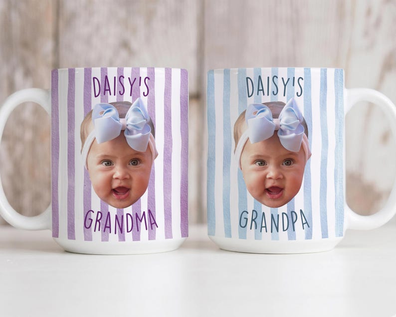 K&ouml;nnte beinhalten: Zwei wei&szlig;e Keramikbecher mit einem Babygesicht und den Texten "DAISY'S GRANDMA" und "DAISY'S GRANDPA". Die Becher haben vertikale Streifenhintergr&uuml;nde in Lila und Blau.