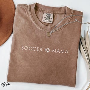 May include: A brown t-shirt with the text "SOCCER MAMA" and a soccer ball graphic printed in white. The shirt is made of a soft, comfortable fabric.