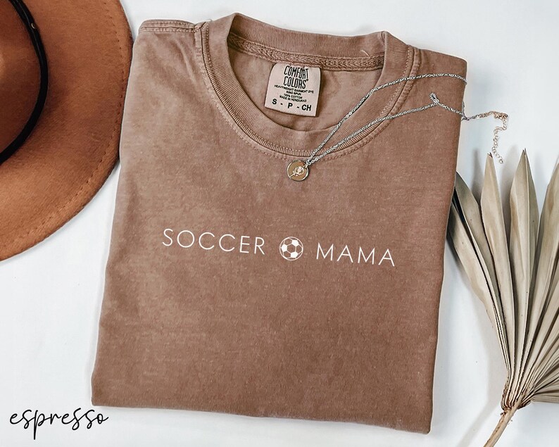 May include: A brown t-shirt with the text "SOCCER MAMA" and a soccer ball graphic printed in white. The shirt is made of a soft, comfortable fabric.