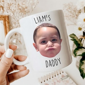 以下が含まれることがあります： 「LIAM'S DADDY」の文字と、むすっとした表情の子供の写真がプリントされた白いセラミックマグカップ。 マグカップにはカーブした持ち手があり、白いマニキュアを塗った手で持たれています。