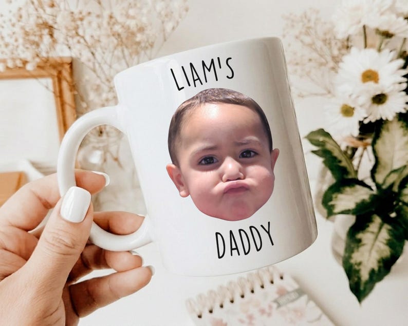以下が含まれることがあります： 「LIAM'S DADDY」の文字と、むすっとした表情の子供の写真がプリントされた白いセラミックマグカップ。 マグカップにはカーブした持ち手があり、白いマニキュアを塗った手で持たれています。