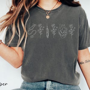 May include: A gray t-shirt with a white outline of a stethoscope, syringe, heart, and flowers. The shirt has a relaxed fit and short sleeves.