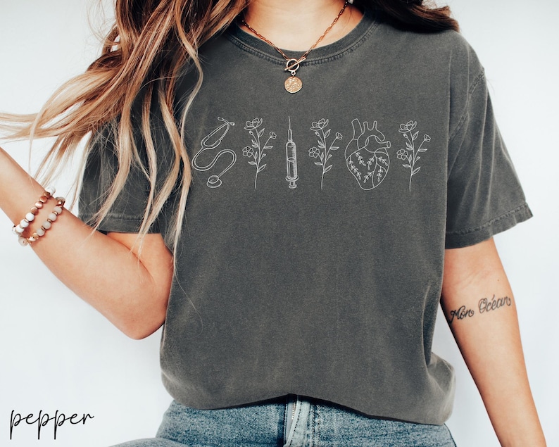 May include: A gray t-shirt with a white outline of a stethoscope, syringe, heart, and flowers. The shirt has a relaxed fit and short sleeves.