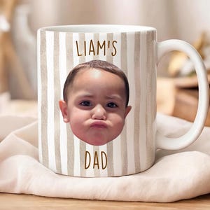 K&ouml;nnte beinhalten: Wei&szlig;e Keramik-Tasse mit Henkel, mit einem Foto eines Babygesichts und dem Text "LIAM'S DAD" in Braun. Die Tasse hat ein vertikales Streifenmuster in Beige und Wei&szlig;.