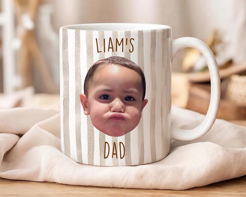K&ouml;nnte beinhalten: Wei&szlig;e Keramik-Tasse mit Henkel, mit einem Foto eines Babygesichts und dem Text "LIAM'S DAD" in Braun. Die Tasse hat ein vertikales Streifenmuster in Beige und Wei&szlig;.