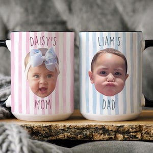 K&ouml;nnte beinhalten: Zwei Keramikbecher mit schwarzen Griffen. Ein Becher zeigt das Gesicht eines Babys mit dem Text "DAISY'S MOM" auf einem rosa-wei&szlig; gestreiften Hintergrund. Der andere Becher hat das Gesicht eines Babys mit dem Text "LIAM'S DAD" auf einem blau-wei&szlig; gestreiften Hintergrund.