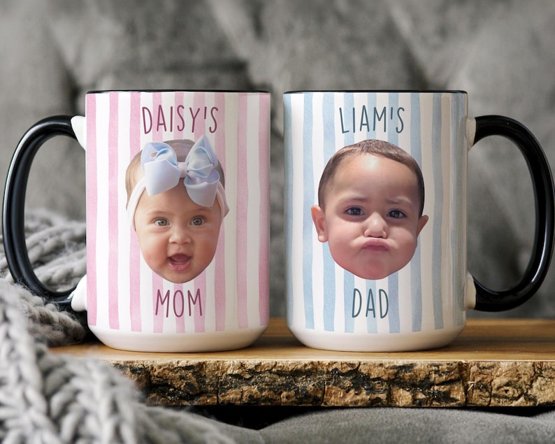 K&ouml;nnte beinhalten: Zwei Keramikbecher mit schwarzen Griffen. Ein Becher zeigt das Gesicht eines Babys mit dem Text "DAISY'S MOM" auf einem rosa-wei&szlig; gestreiften Hintergrund. Der andere Becher hat das Gesicht eines Babys mit dem Text "LIAM'S DAD" auf einem blau-wei&szlig; gestreiften Hintergrund.