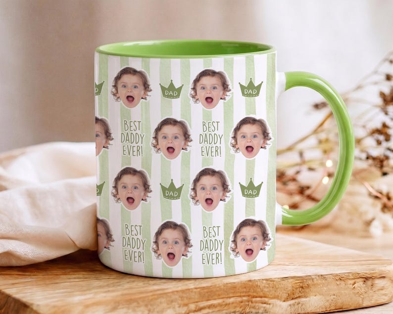 Może przedstawiać: Biały ceramiczny kubek z zielonym wnętrzem i uchwytem. Kubek ma powtarzający się wz&oacute;r twarzy dziecka, koron i tekstu "BEST DADDY EVER!" i "DAD" na paskowanym tle.