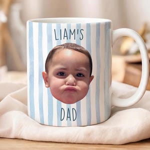 K&ouml;nnte beinhalten: Wei&szlig;e Keramik-Tasse mit blau-wei&szlig;em Streifenmuster. Die Tasse zeigt das Gesicht eines Kindes und den Text "LIAM'S DAD" in Blau. Sie hat einen wei&szlig;en Henkel und liegt auf einem wei&szlig;en Tuch.