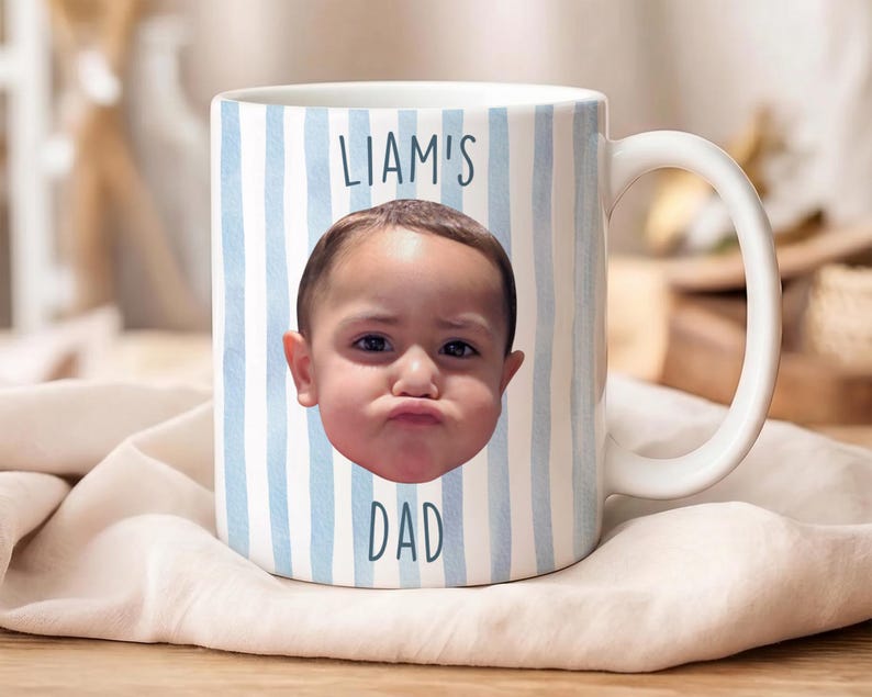 K&ouml;nnte beinhalten: Wei&szlig;e Keramik-Tasse mit blau-wei&szlig;em Streifenmuster. Die Tasse zeigt das Gesicht eines Kindes und den Text "LIAM'S DAD" in Blau. Sie hat einen wei&szlig;en Henkel und liegt auf einem wei&szlig;en Tuch.