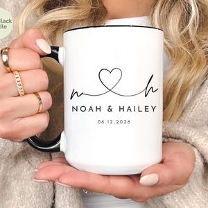 May include: White ceramic mug with a black handle and rim. The mug features a heart-shaped design with the names "Noah & Hailey" and the date "06.12.2026" printed in black. The mug is labeled "15oz Black Handle".