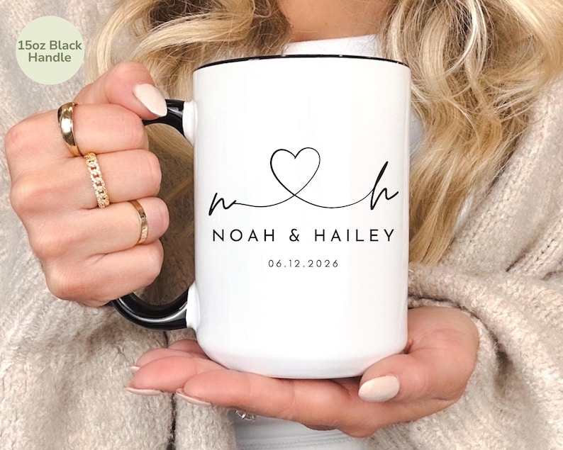 May include: White ceramic mug with a black handle and rim. The mug features a heart-shaped design with the names "Noah & Hailey" and the date "06.12.2026" printed in black. The mug is labeled "15oz Black Handle".