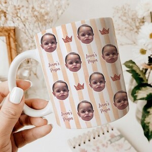 Może przedstawiać: Biały ceramiczny kubek z uchwytem, ​​z powtarzającym się wzorem twarzy dziecka, koron i napisem "Liam's Papa" na paskowanym tle. Kubek jest trzymany przez osobę.